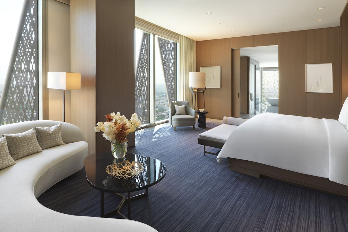 Une chambre d’hôtel moderne avec un canapé incurvé, un lit, une petite table sur laquelle est posé un vase de fleurs, et une vue dégagée grâce à de grandes fenêtres.