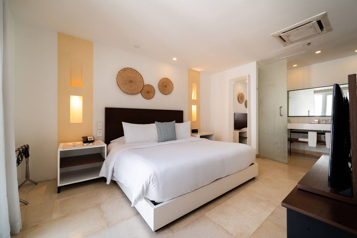 Habitación de hotel moderna con una cama grande, mesitas de noche, un televisor de pantalla plana y un baño con espejo y lavamanos.
