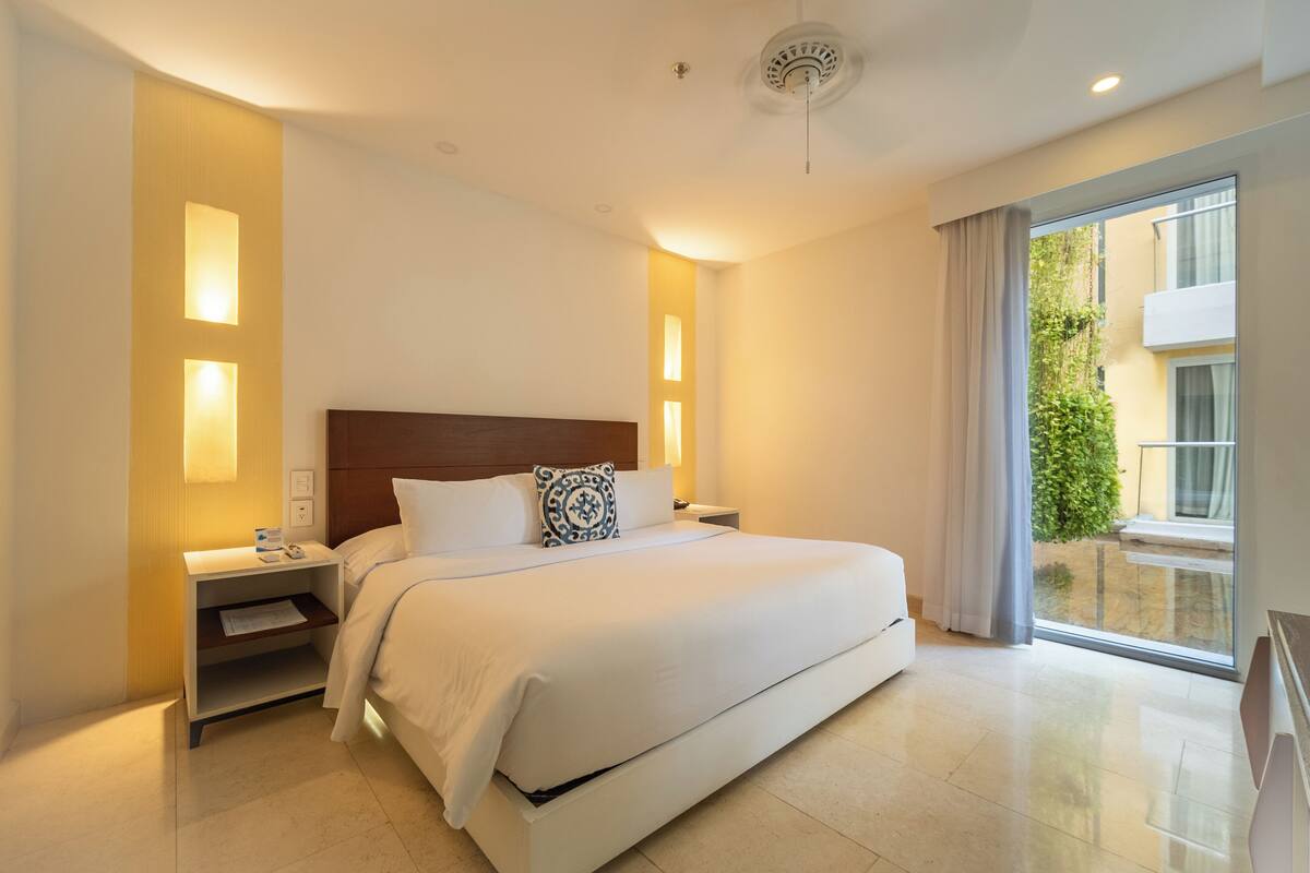 Una habitación de hotel moderna con una cama grande, mesitas de noche y vistas al exterior a través de un amplio ventanal.
