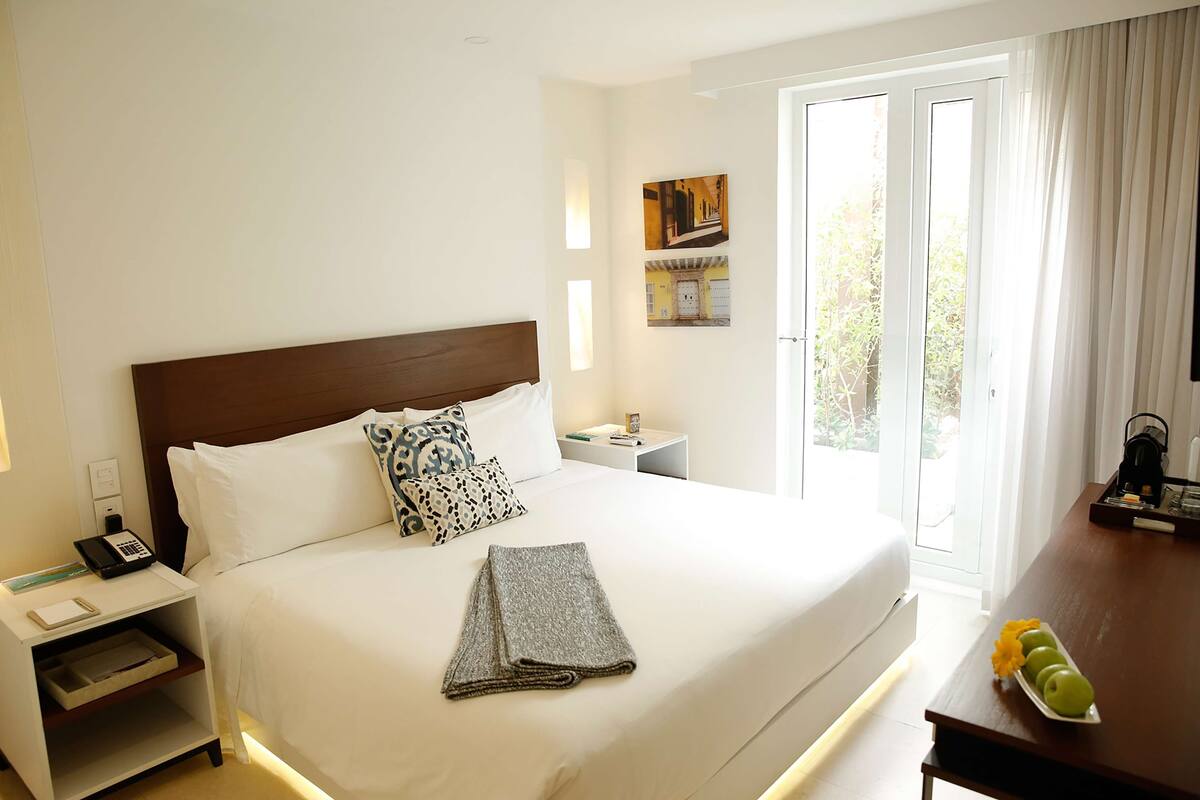 Habitación de hotel con una cama grande, una mesita de noche con teléfono, cortinas y vistas al exterior con vegetación.