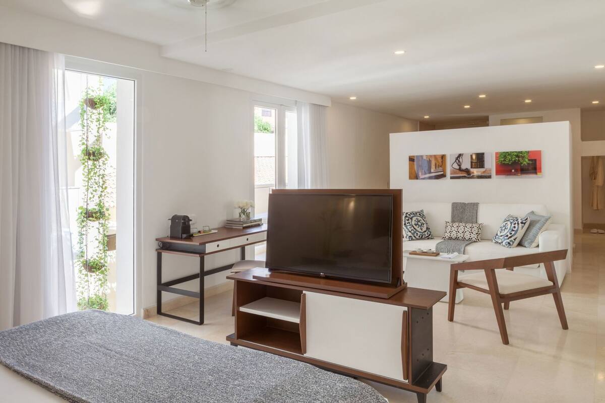 Een moderne woonkamer met een flatscreen televisie op een houten standaard, een bureau met een koffiezetapparaat, een bank met gekleurde kussens en een groot raam met uitzicht op groen.