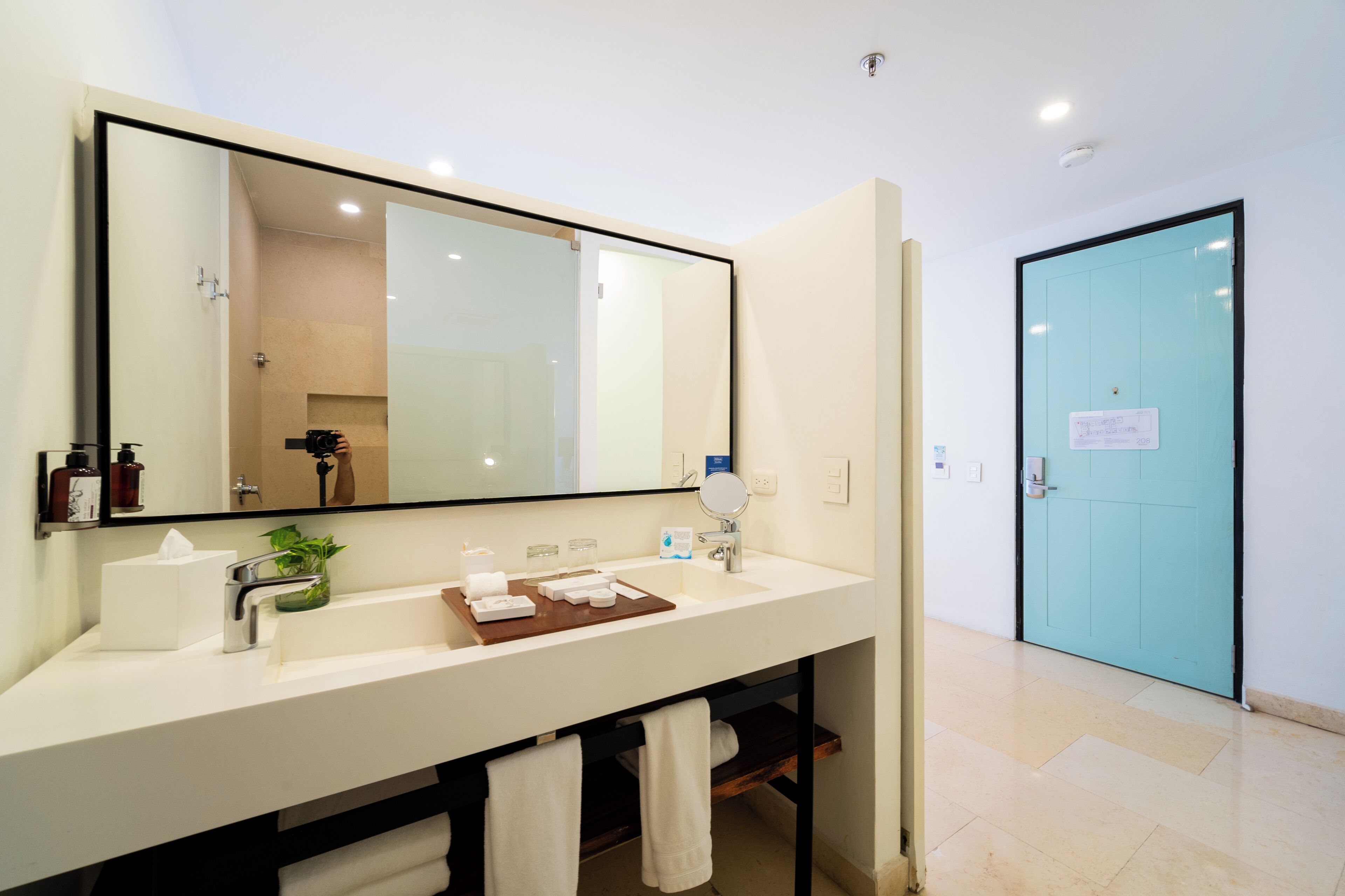 Un baño moderno con un espejo grande, lavamanos blanco y una bandeja de madera. Se ve un perchero y una puerta con un letrero.