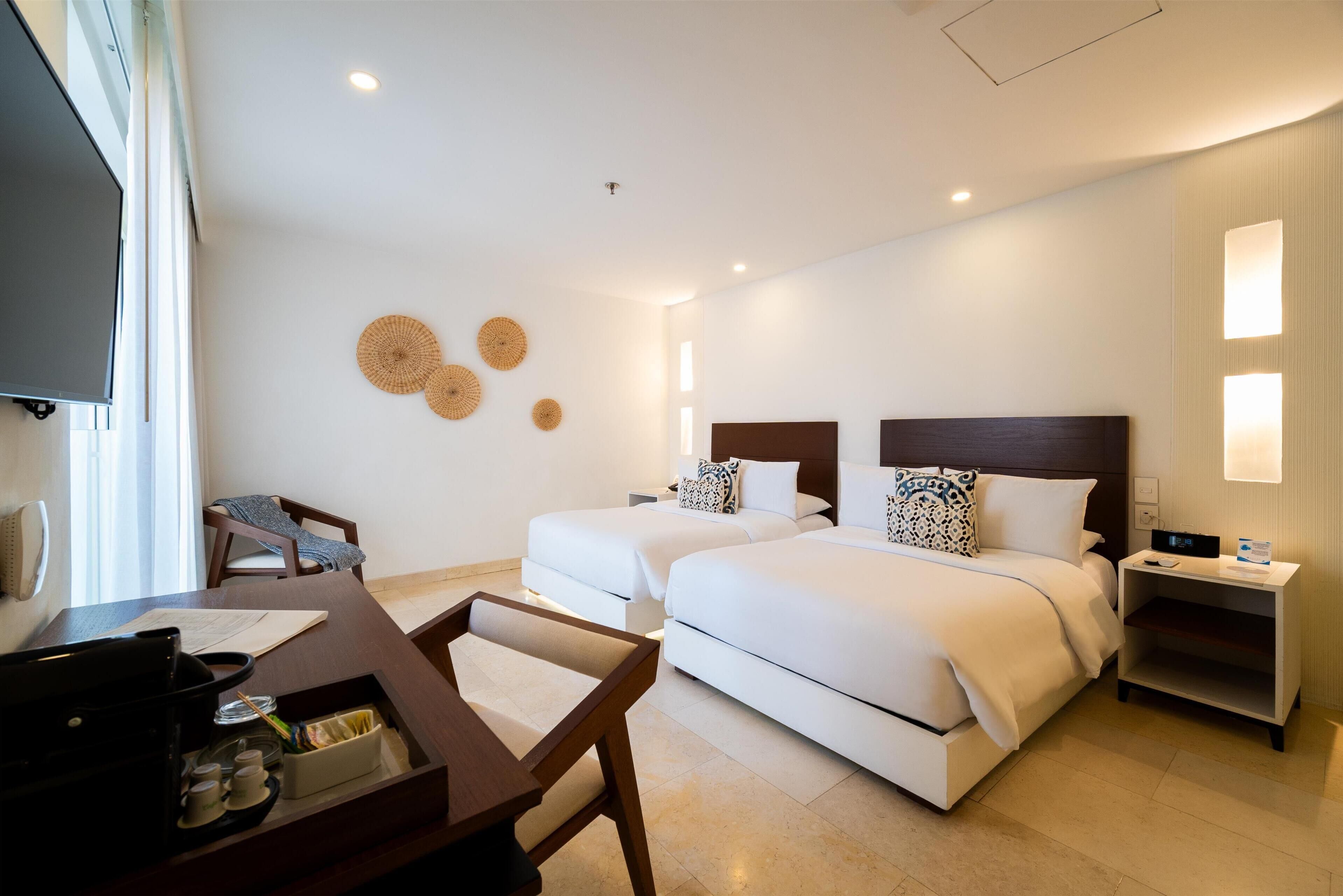 Habitación de hotel con dos camas, un escritorio con silla, televisor y mesita de noche con lámpara.