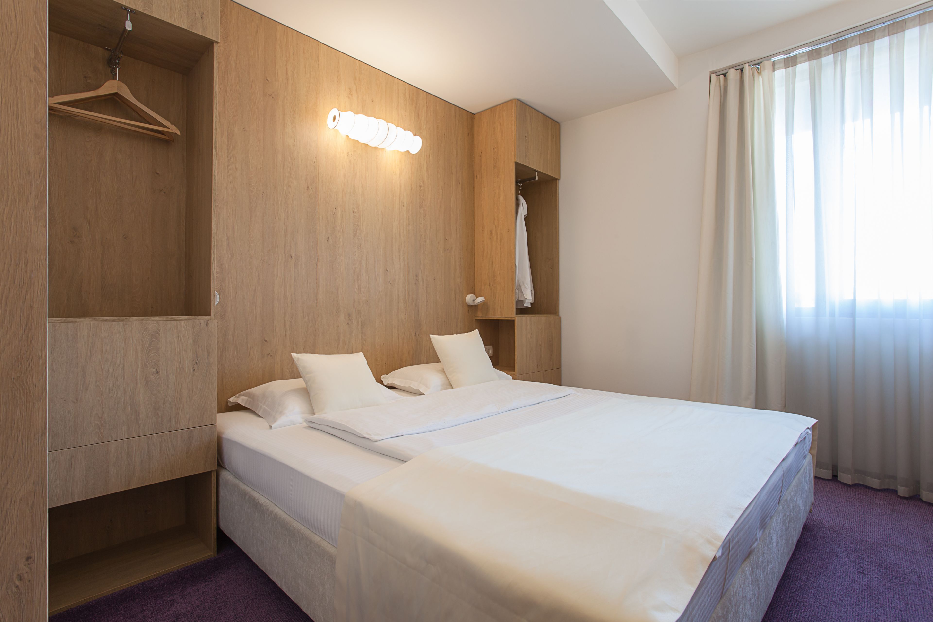 Habitación de hotel con una cama grande, paredes de paneles de madera, un perchero y una ventana con cortinas.