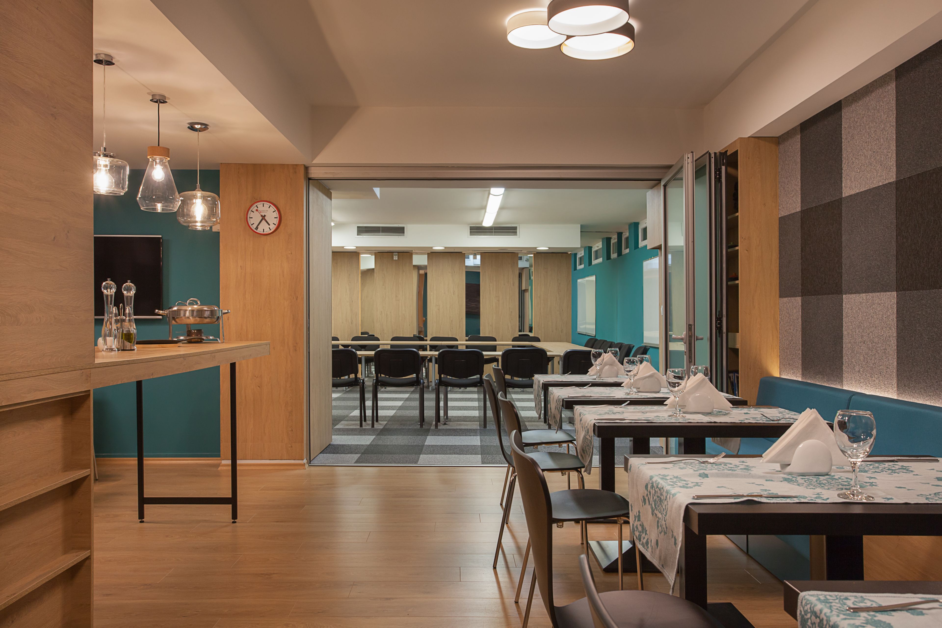 A modern dining area with wooden tables and chairs, a kitchen counter with glassware, and a view into a dining hall.