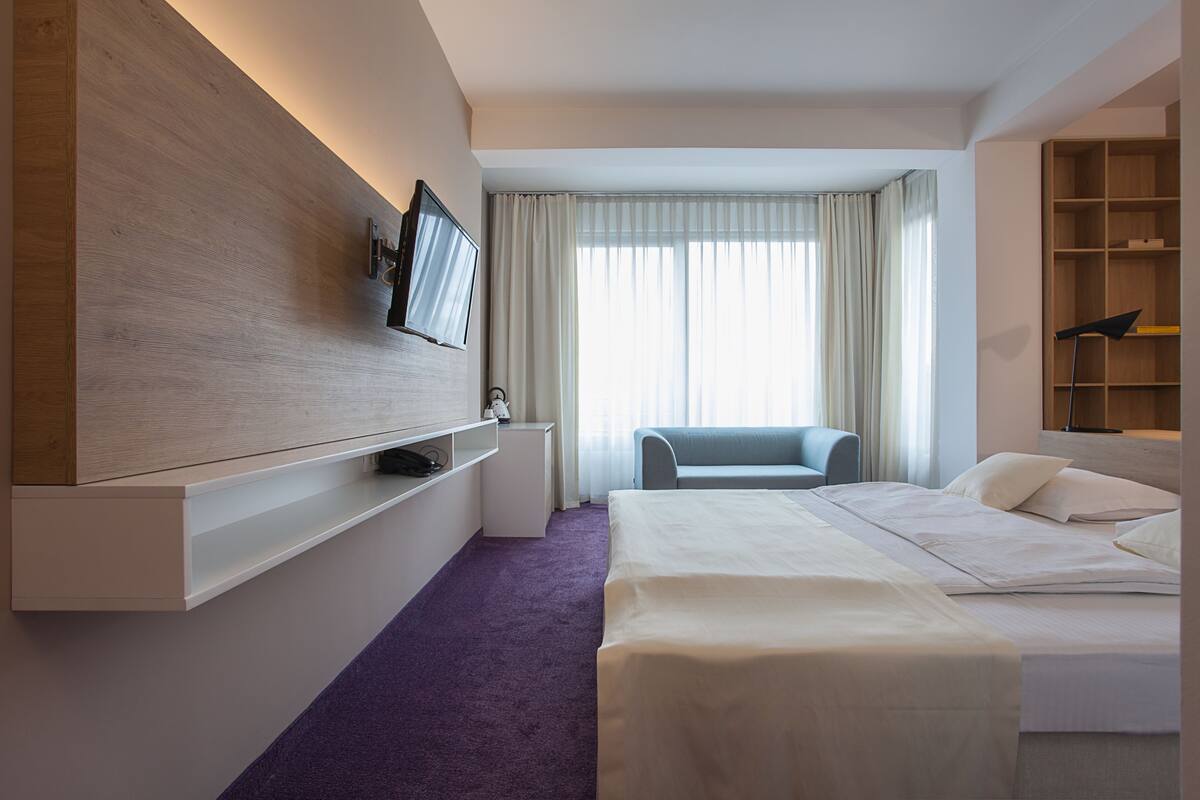 Ein modernes Hotelzimmer mit einem Bett, einem an einer Holzplatte montierten Fernseher, einem kleinen Schreibtisch und einem Sessel.