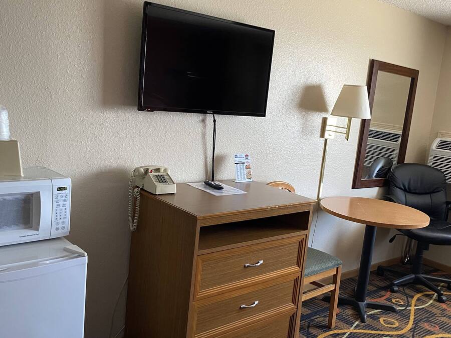 Habitación de hotel con televisor de pantalla plana en la pared, microondas, teléfono, una pequeña mesa con una silla, escritorio con cajones, lámpara, aire acondicionado y alfombra estampada.