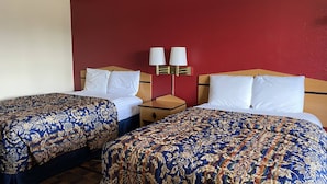 Une chambre d’hôtel avec deux lits simples, chacun recouvert d’une couvre-lit à motifs floraux, une table de chevet en bois avec une lampe, et un mur d’accent rouge.