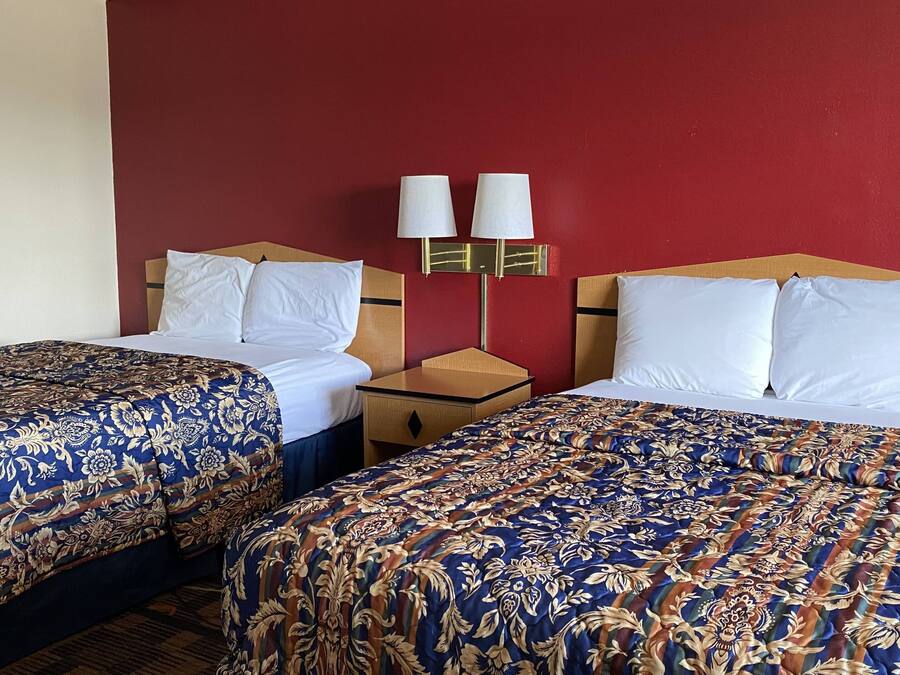 A hotel room with two single beds, each with a floral-patterned bedspread, a wooden nightstand with a lamp, and a red accent wall.