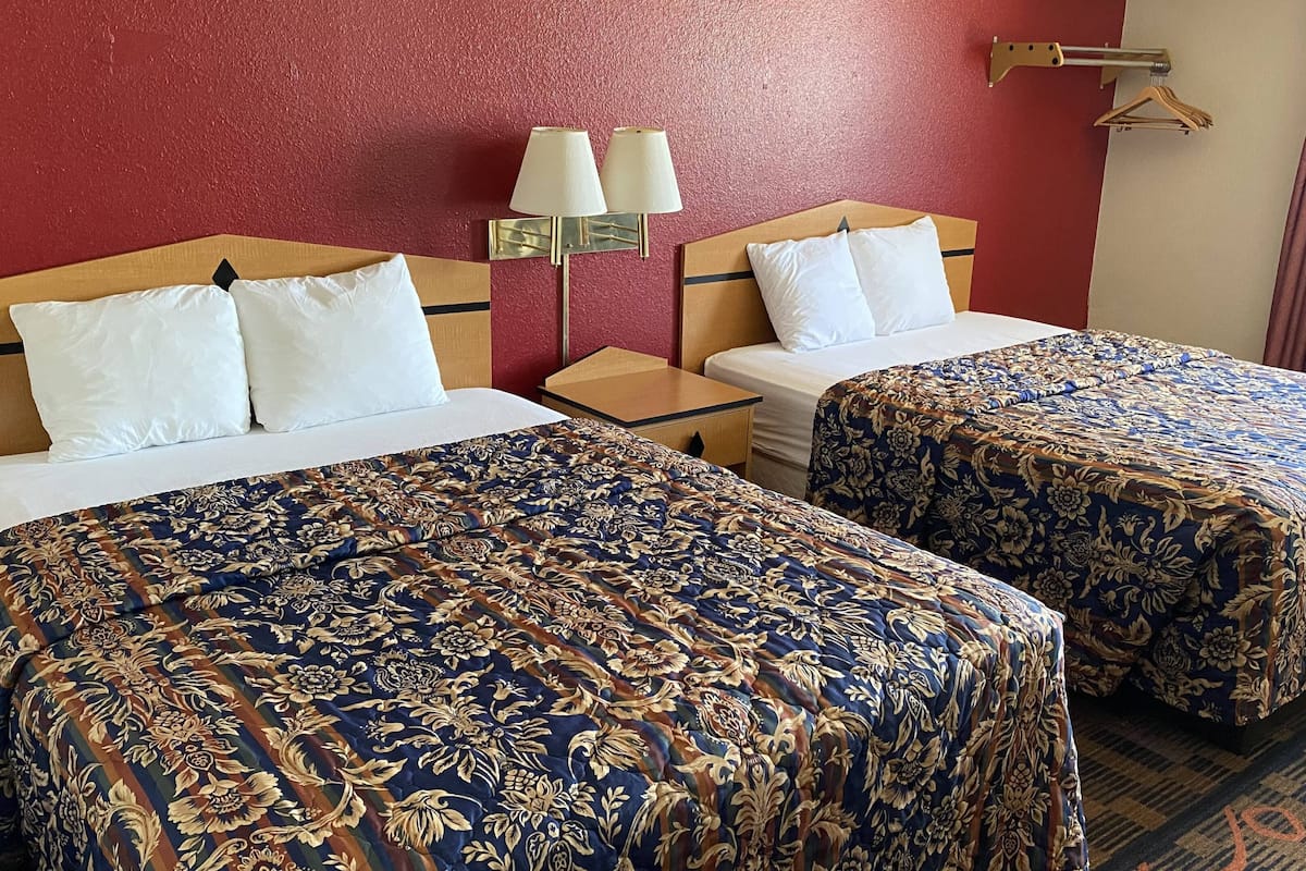 Habitación de hotel con dos camas, cada una con una colcha de motivos florales, una mesita de noche con lámpara y una pared roja.