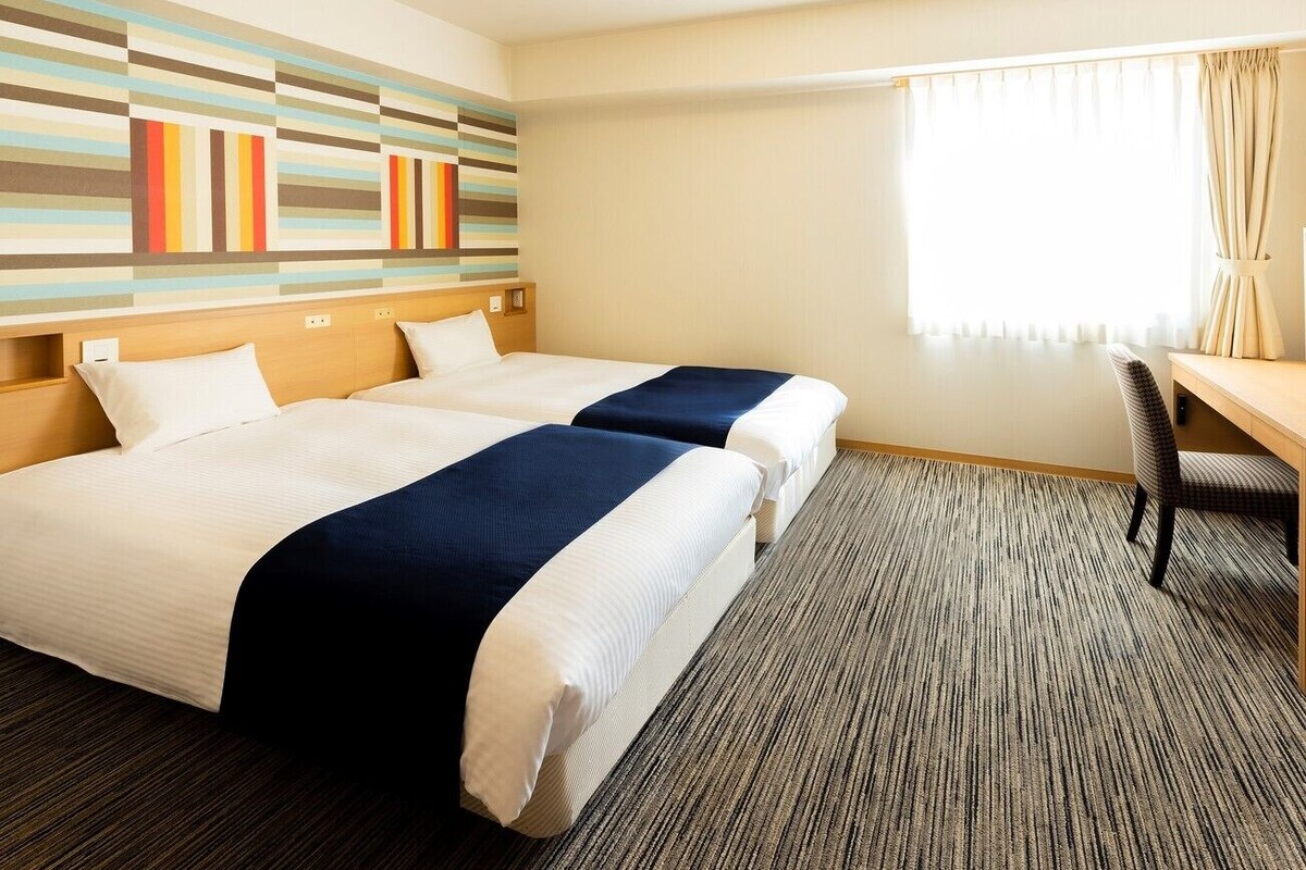 A hotel room with two beds, a desk, a chair, and a striped wall decoration.