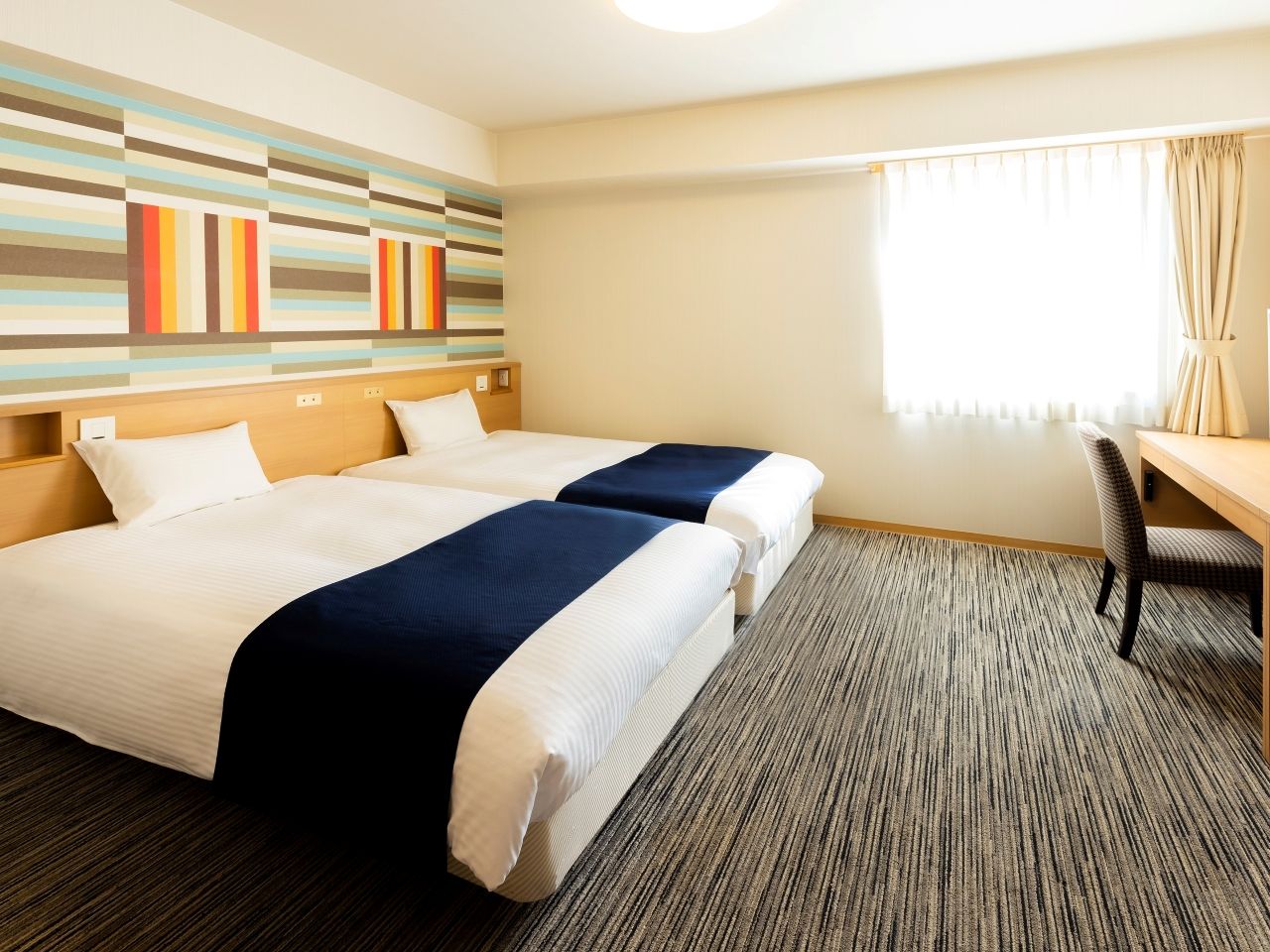 Quarto de hotel com duas camas, uma escrivaninha e janela com cortinas.