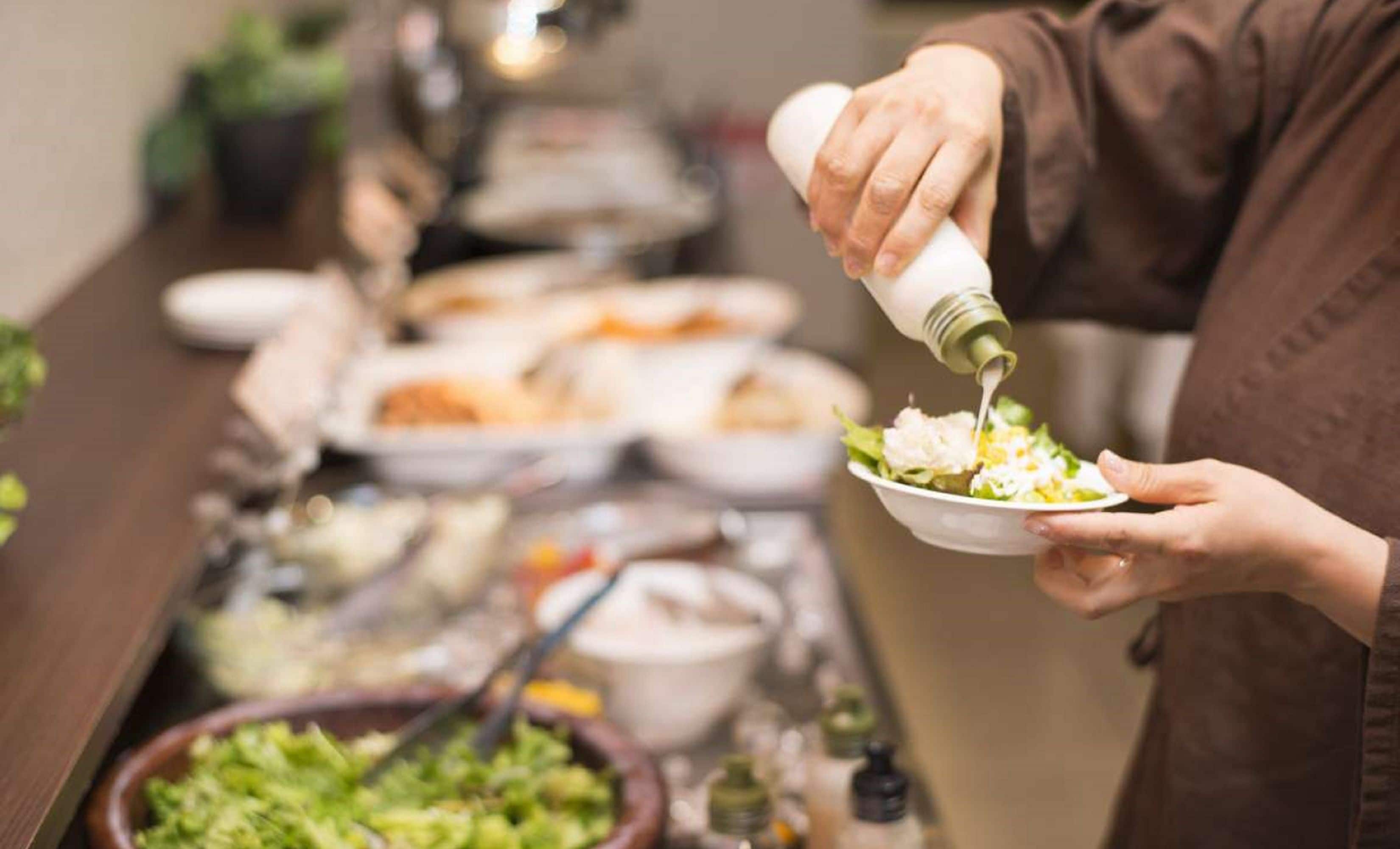 Un buffet proposant divers plats, et une personne qui verse de la vinaigrette sur une salade.
