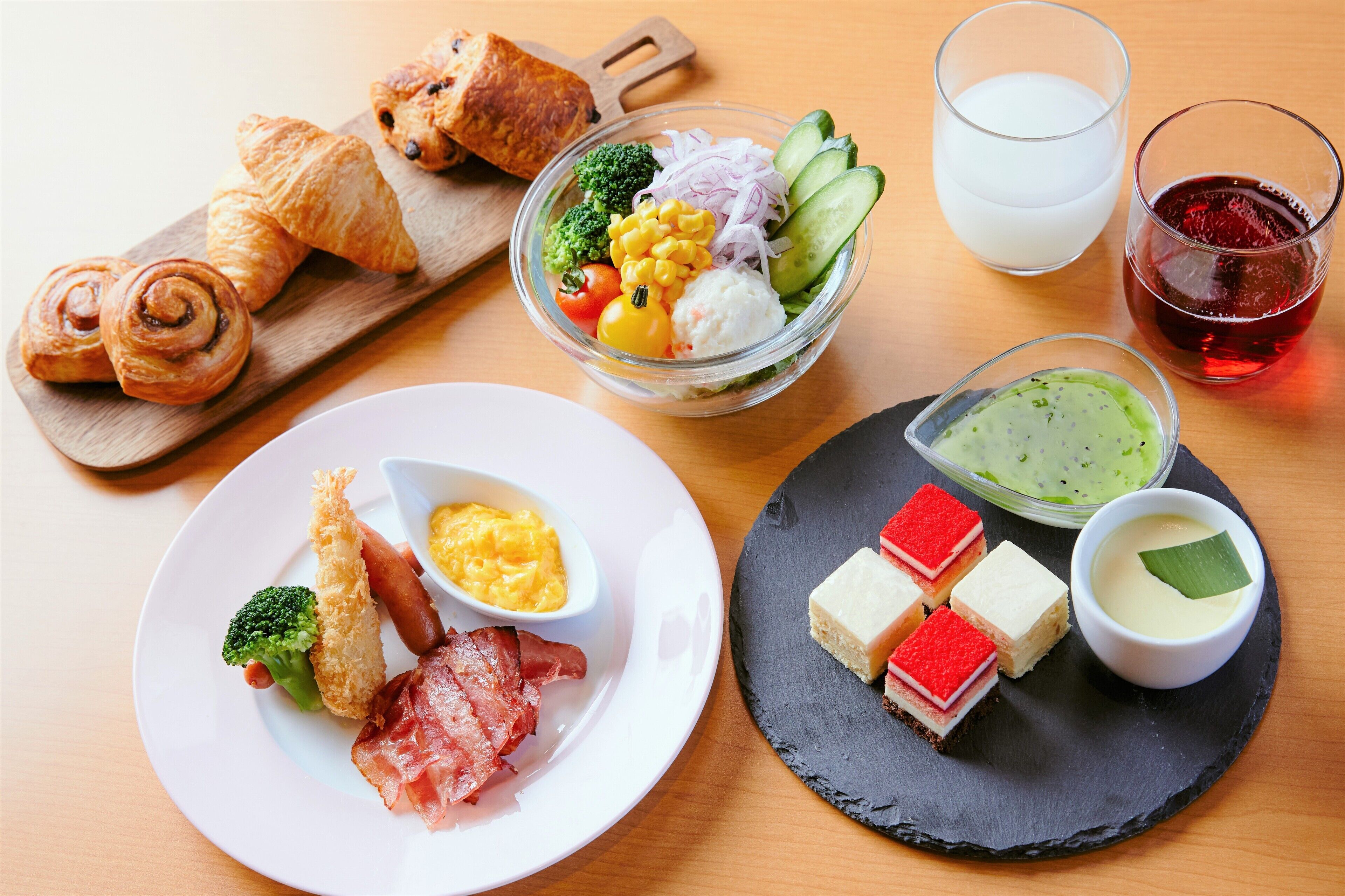 A breakfast spread with croissants, a fruit and vegetable bowl, a glass of milk, a glass of red drink, a plate with fried food and eggs, and a slate board with desserts.