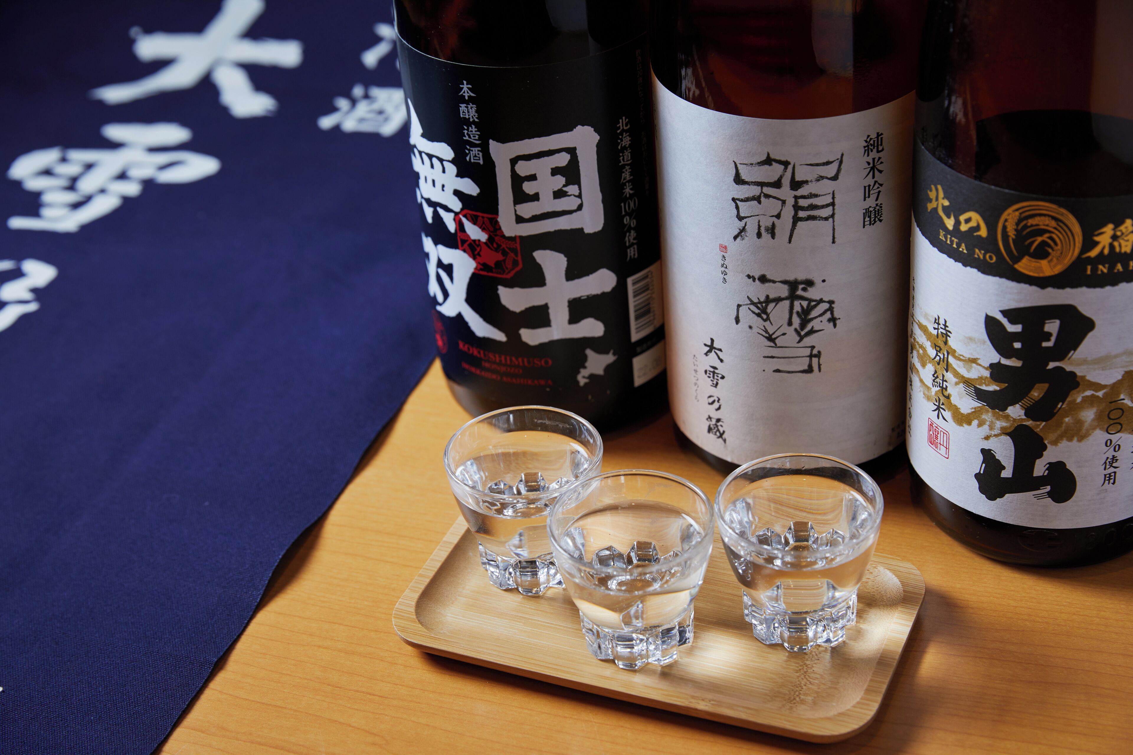 Three bottles of sake with Japanese labels, placed on a wooden tray with shot glasses filled with water.