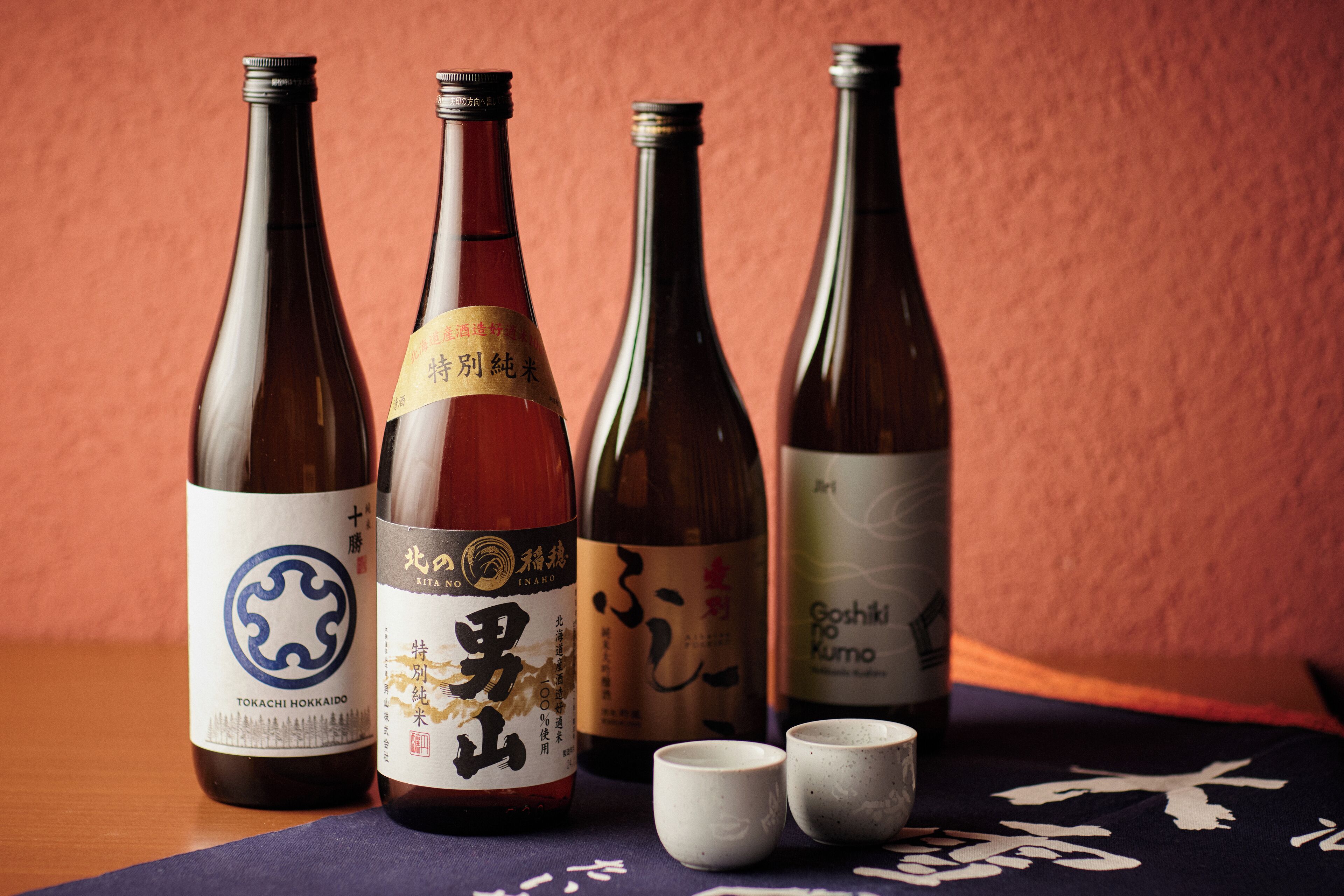 Four bottles of Japanese sake with labels, two small cups, and a tablecloth with a pattern.