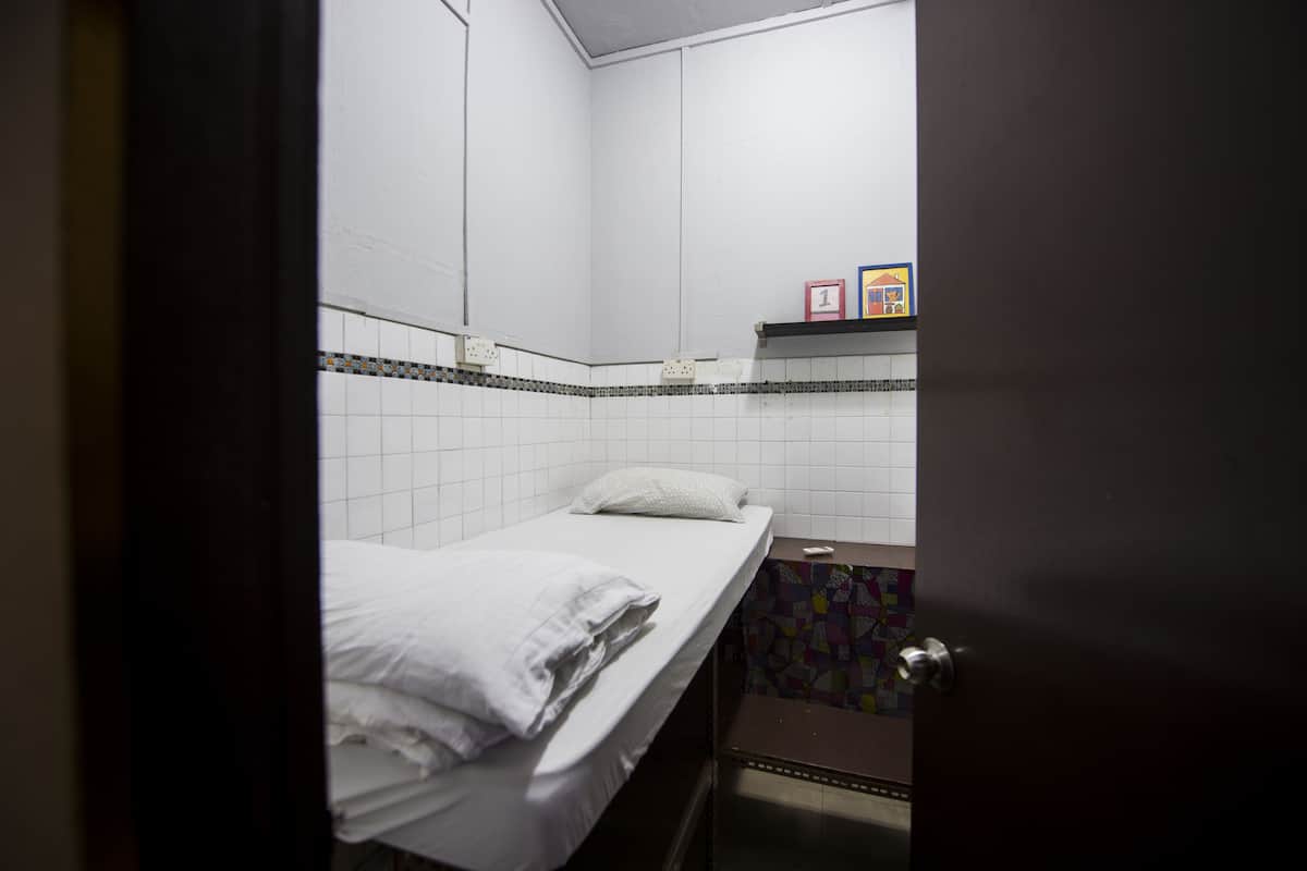 Une petite chambre avec un lit simple, du linge de lit blanc, un mur carrelé, une petite étagère avec des blocs numérotés et une porte.