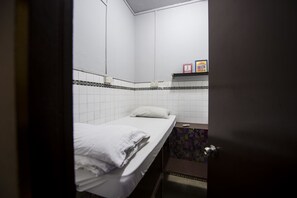 A small, single-bed room with white bedding, a tiled wall, a small shelf with numbered blocks, and a door. - Urban Hostel (Singapore)