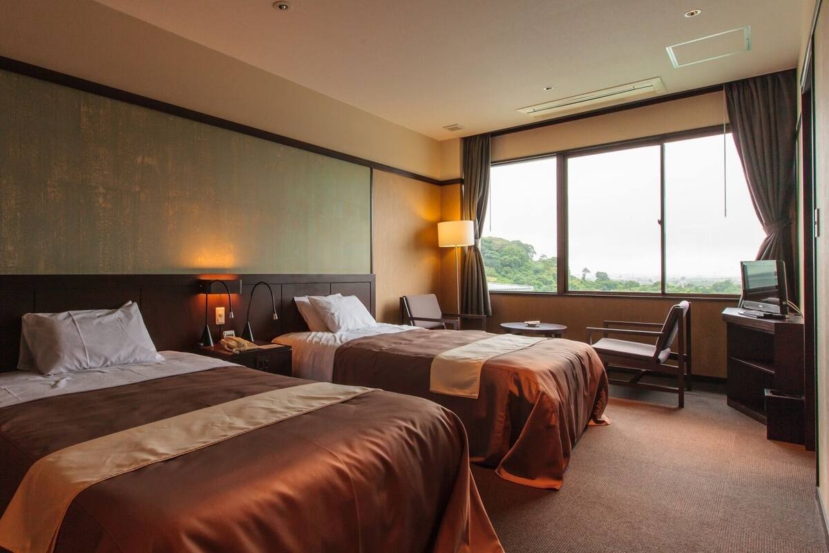 A hotel room with two beds, a desk, a chair, a TV, and a large window with a view.