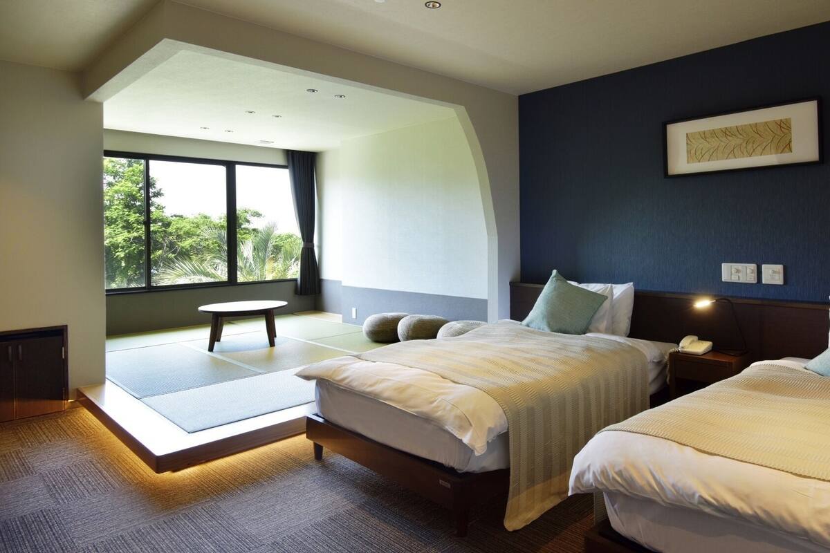 A modern hotel room with two beds, a large window with a view, a small table, and a ceiling light.