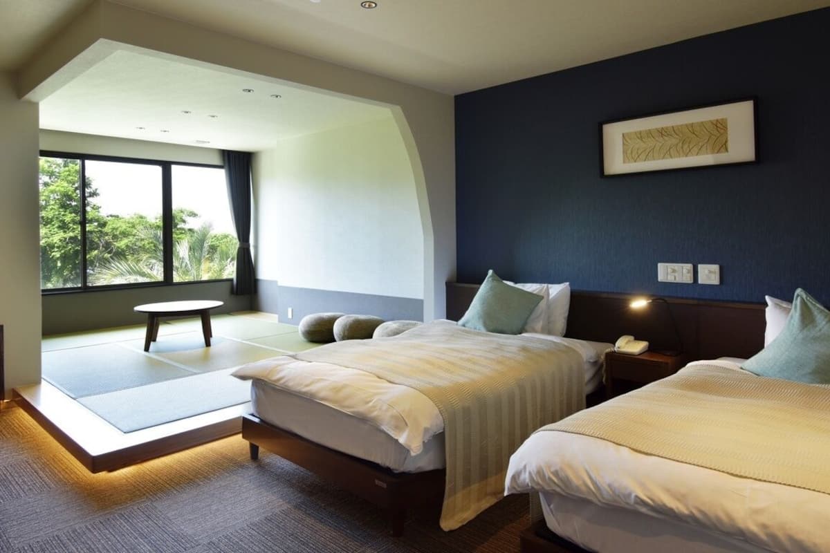 A modern hotel room with two beds, a large window, and a view of greenery.