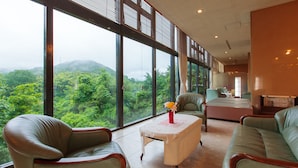 Amplia habitación con ventanas grandes que ofrecen vistas a exuberante vegetación y colinas. Hay una mesa de comedor con sillas y un sofá.