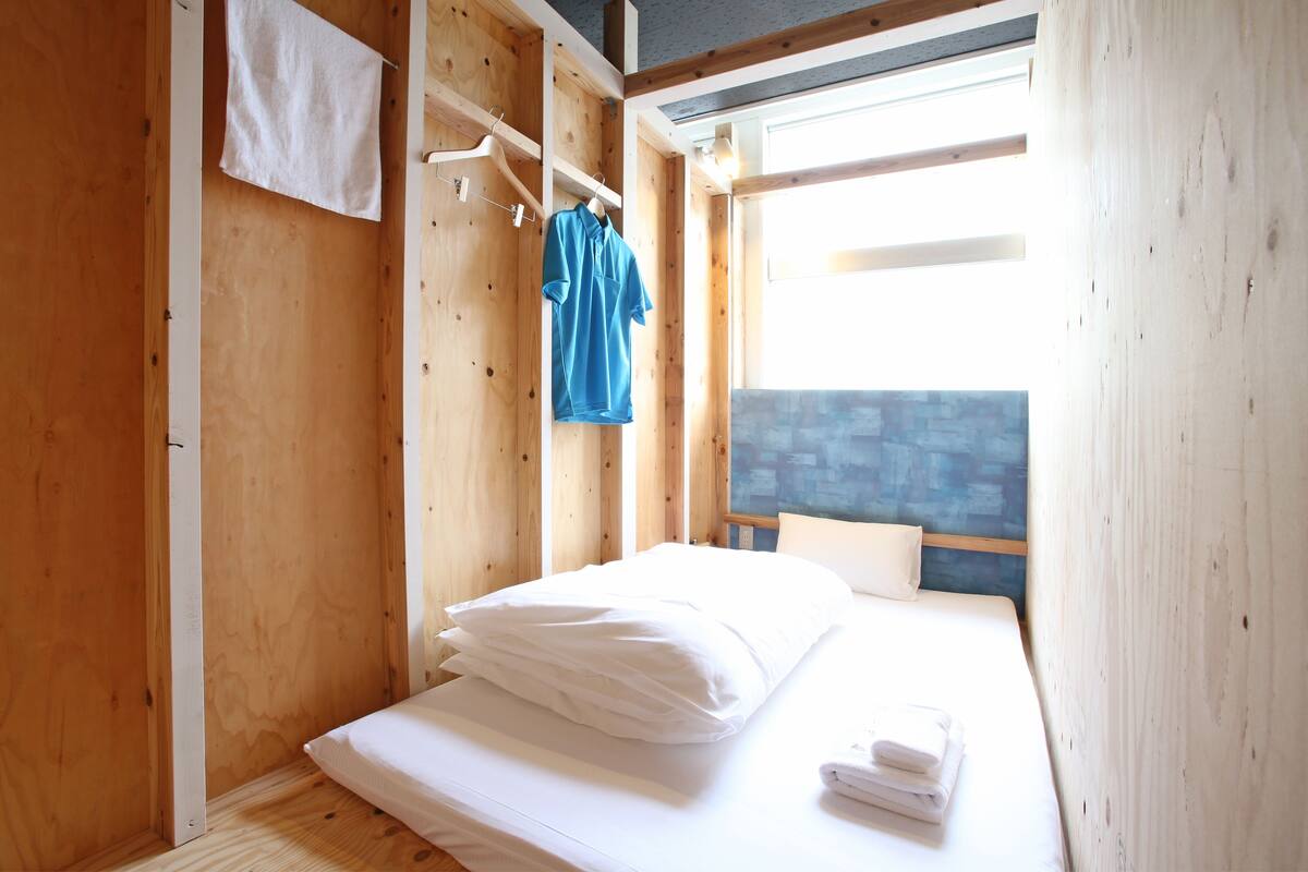 Ein kleiner, hölzerner Raum mit einem Bett, einem Haken für Handtücher und einem Fenster.