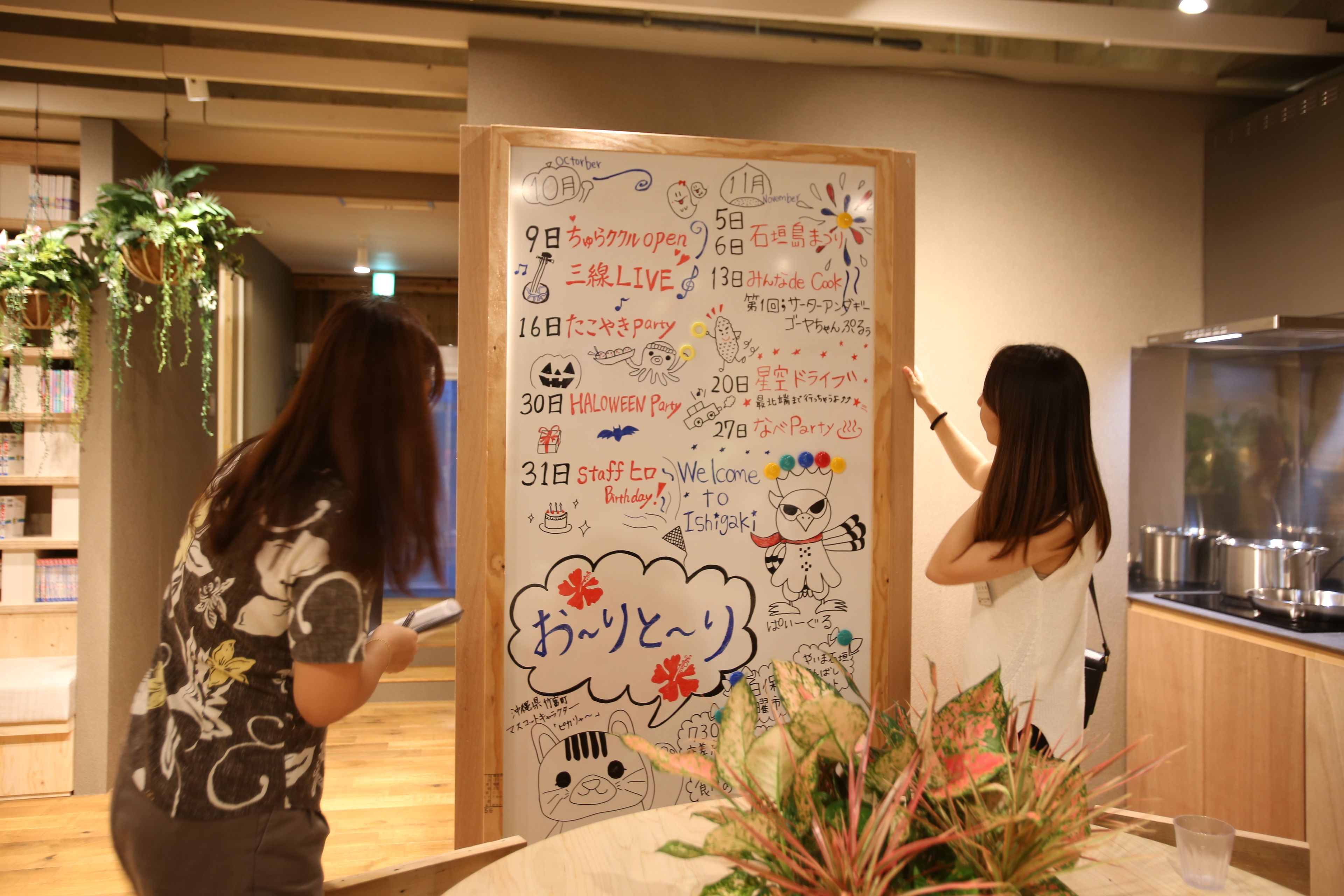 Un tableau blanc sur lequel sont inscrits en japonais les dates et les noms des événements, entouré de plantes et d’un coin cuisine.