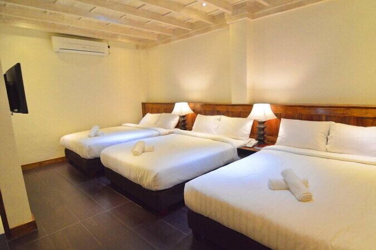 A hotel room with three single beds, each with a wooden headboard and white bedding. There is a bedside table with a lamp on each bed.