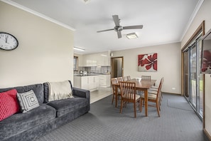 Ein Wohnzimmer mit grauem Sofa, einem Esstisch mit Stühlen, einem Deckenventilator und einer Küche im Hintergrund.