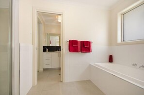 A modern bathroom with a white bathtub, a glass shower enclosure, and a visible kitchen area in the background. - Fringe Apartments (Mount Gambier)