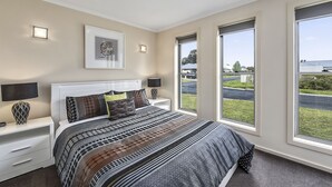 A bedroom with a large bed, two bedside tables with lamps, a framed artwork above the bed, and a view of a road and greenery outside. - Megan Court Apartments (Mount Gambier)