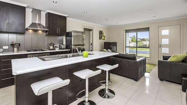 A modern kitchen with dark cabinetry, a white countertop, and a central island with bar stools. - Megan Court Apartments (Mount Gambier)
