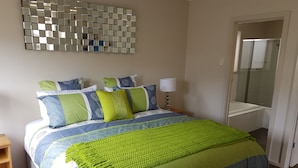A neatly made bed with a green and blue bedspread, a mirror on the wall, and a door leading to a bathroom. - Hart Street Apartment (Mount Gambier)