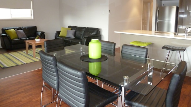 A modern living room with a glass dining table, black leather chairs, a black leather sofa, and a small wooden coffee table. - Hart Street Apartment (Mount Gambier)