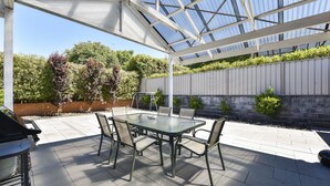 Eine Gartenlaube mit einem Glastisch und Stühlen, umgeben von einem Garten und einer Ziegelmauer.