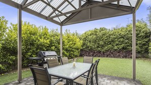 An outdoor patio with a glass table and chairs, a grill, and a covered area with a metal roof. - MG Delux Apartment (Mount Gambier)