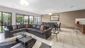 A modern living room with a sectional sofa, a glass coffee table, and a dining area with chairs and a table. - MG Delux Apartment (Mount Gambier)