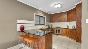 A modern kitchen with a central island, bar stools, wooden cabinets, and a window with blinds. - MG Delux Apartment (Mount Gambier)