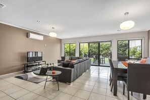 A modern living room with a sofa, TV, dining table, and chairs. - MG Delux Apartment (Mount Gambier)