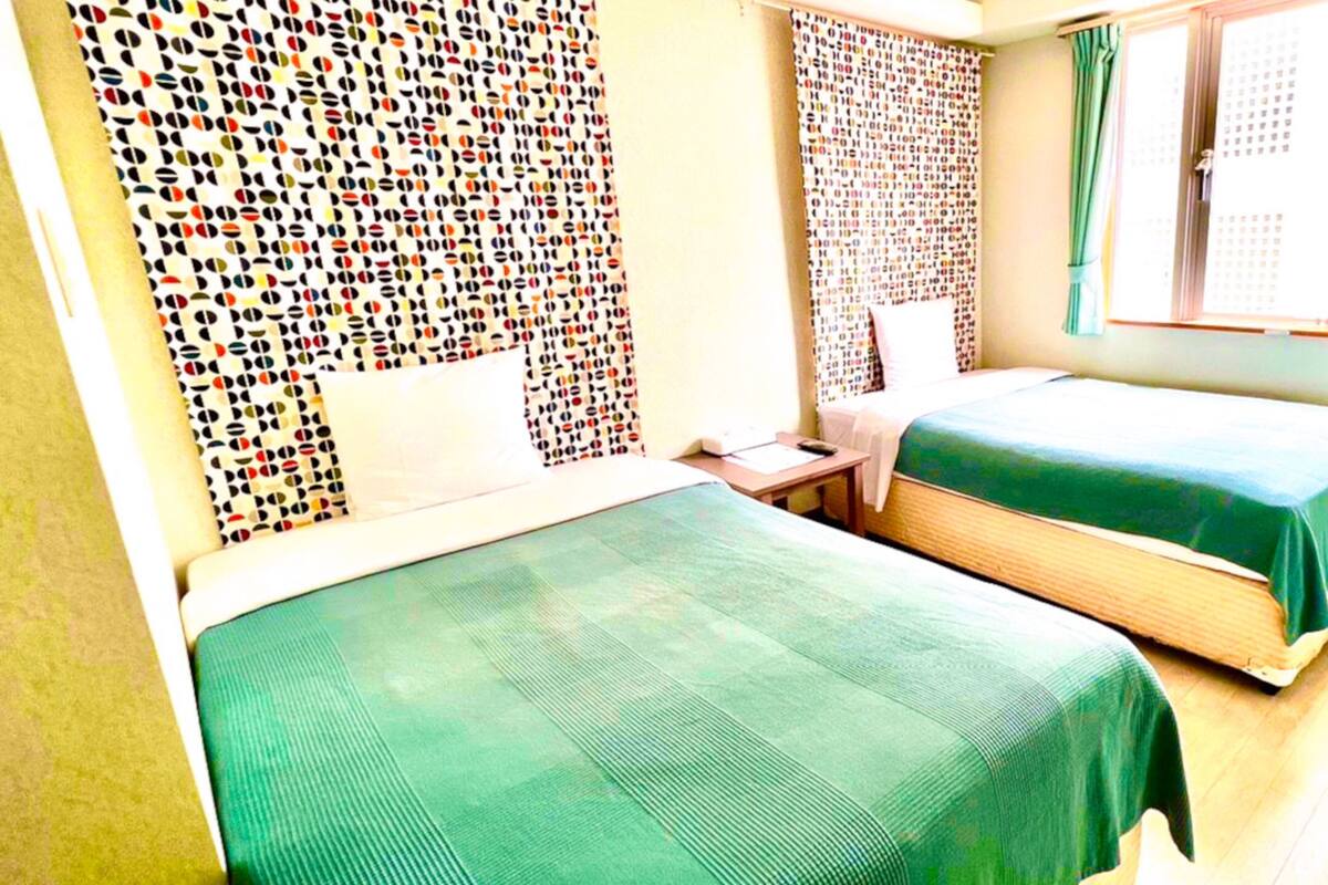 Una habitación con dos camas, una mesita, una ventana con cortinas y un diseño estampado en la pared.