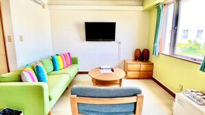 A small, well-lit room with a green sofa, a wooden chair, a small table, a TV mounted on the wall, and a window with curtains. - Hotel Happy Holiday Ishigakijima (Ishigaki)