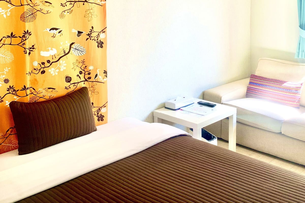 Habitación de hotel con cama, sofá, una mesita con un control remoto y ventana con vista a edificios.