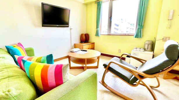A room with a green sofa, a colorful striped pillow, a wooden side table, a small round table, a chair, a television mounted on the wall, and a window with curtains. - Hotel Happy Holiday Ishigakijima (Ishigaki)