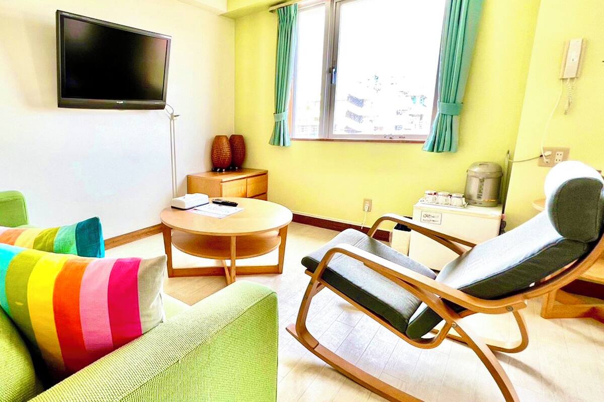 Una habitación con un sofá verde, un cojín a rayas de colores, una mesita de noche de madera, una pequeña mesa redonda, una silla, un televisor montado en la pared y una ventana con cortinas.