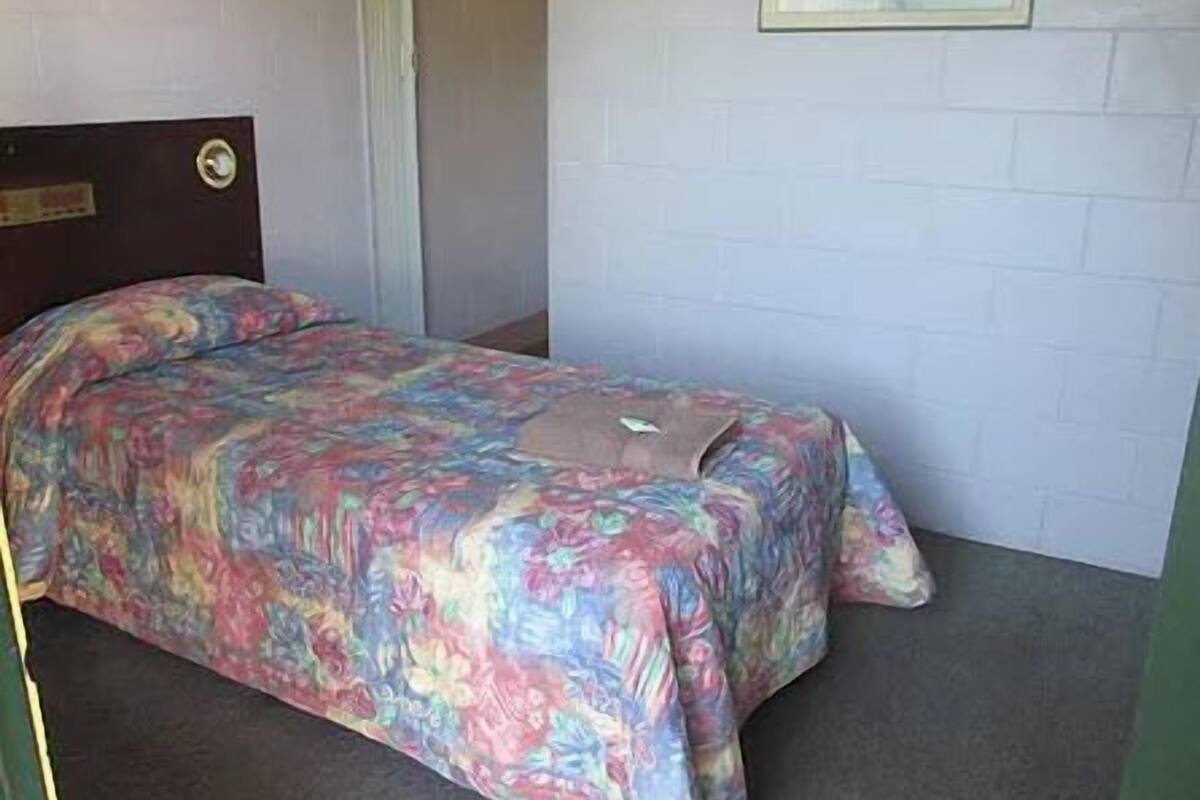 Ein kleines Zimmer mit einem Einzelbett, einer Bettdecke mit Blumenmuster, einem Holz-Kopfteil und einem gerahmten Bild an der Wand.