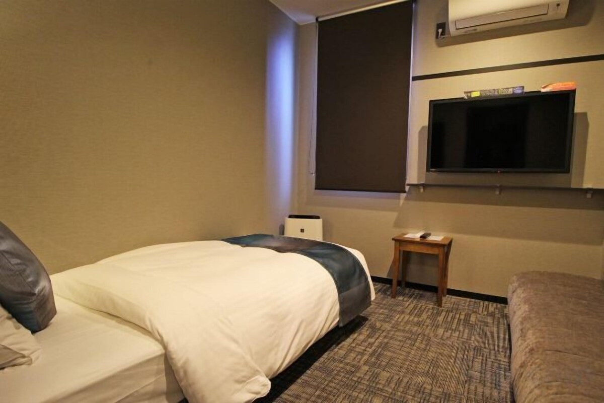 A hotel room with a bed, a small table, a TV mounted on the wall, and a window.