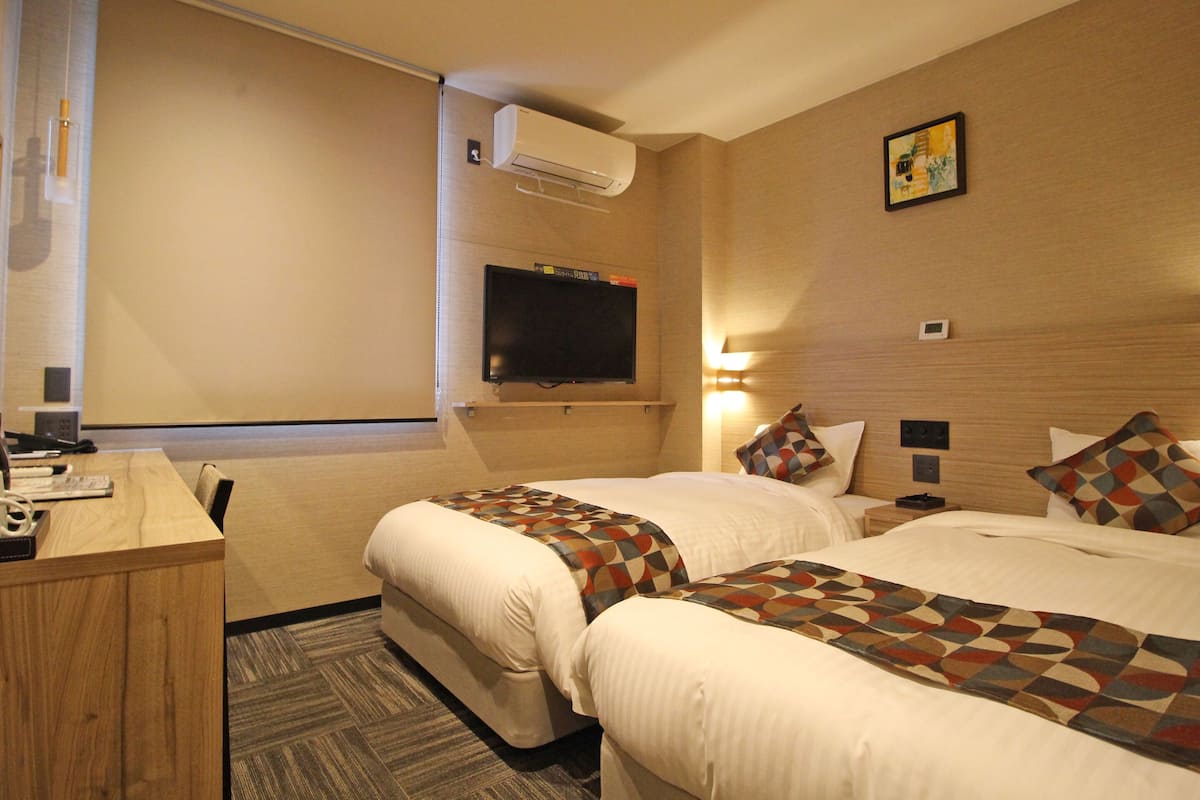 A hotel room with two beds, a TV, a desk, and a wall-mounted air conditioner.