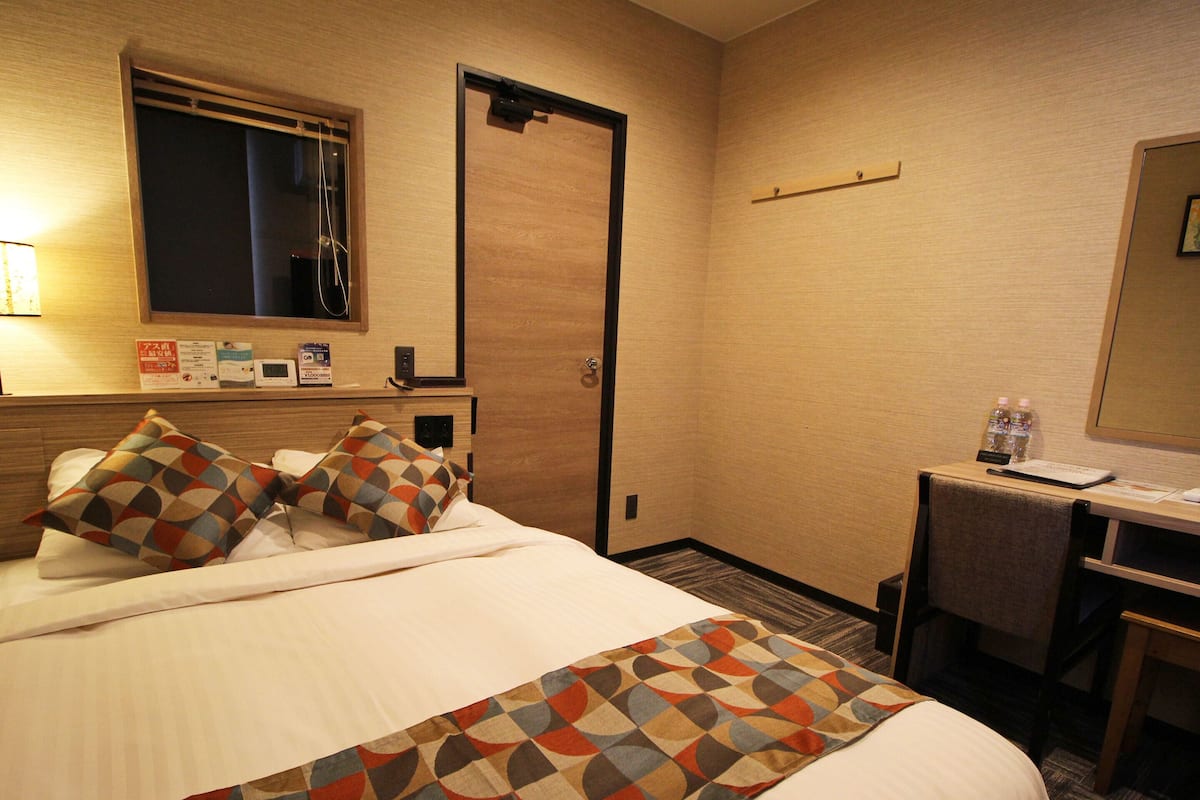 A hotel room with a bed, a desk, a chair, a desk lamp, a mirror, a door, and a window.
