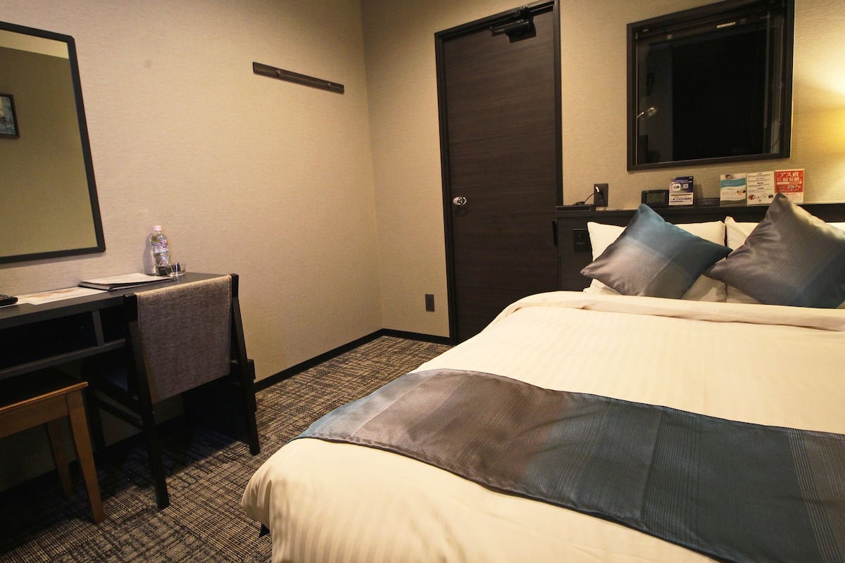 A hotel room with a bed, a desk, a chair, a water bottle, and a door.